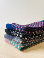 Four pairs of jacquard pattern crew socks in black, navy, brown, and charcoal gray arranged on a wooden table.
